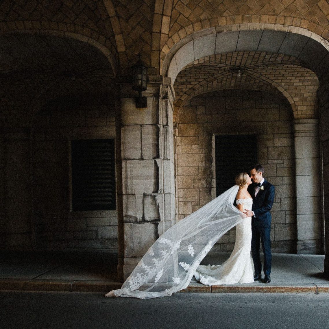 Bride and groom embrace each other under a bow, with a long veil floating.